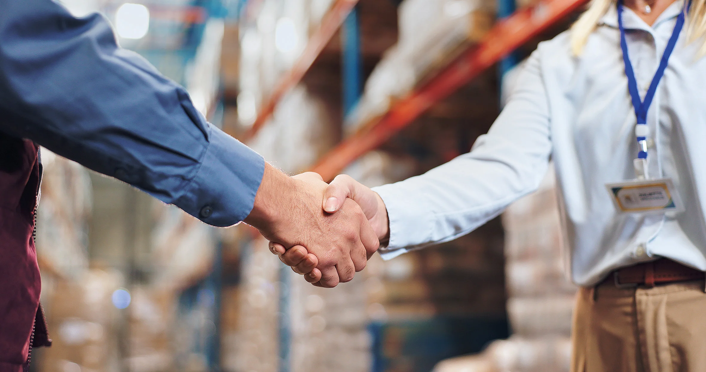 两个人在仓库握手，一个穿蓝色衬衫，另一个穿白色衬衫并佩戴名牌。