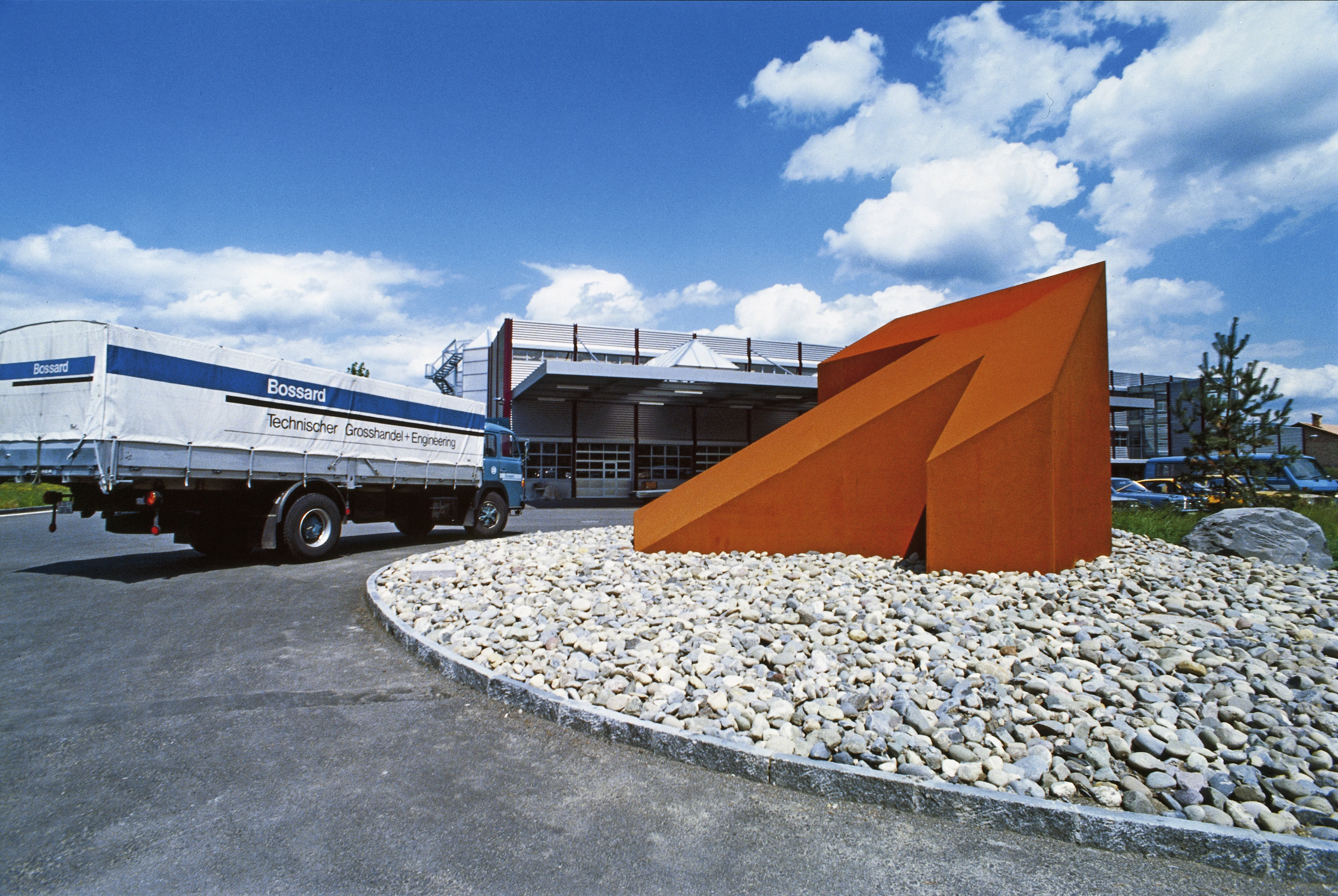 历史图片:一个大型橙色箭头雕塑位于环岛,背景中有Bossard柏中的仓库一辆卡车。