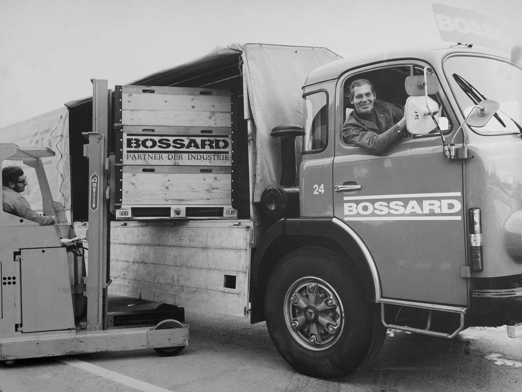 一张过去的黑白照片显示一名男子在堆垛机上卸载Bossard卡车，卡车内的另一名男子微笑，营造出合作的场景。