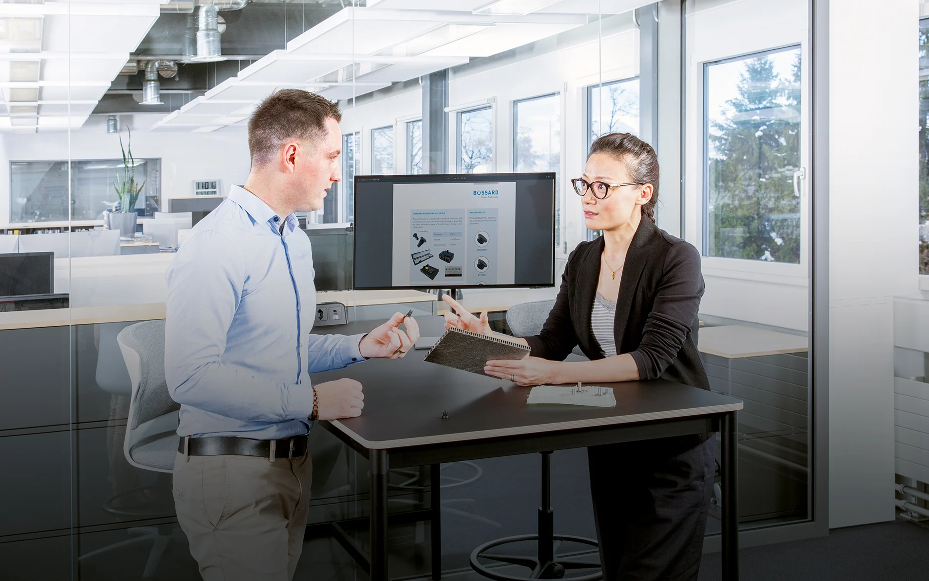 
两个人在现代办公室的桌子上讨论复合材料的紧固问题；一个人拿着夹层板，另一个人拿着紧固件。