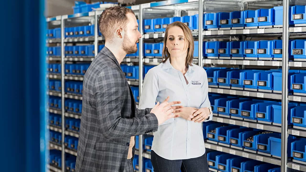 一名男子和一名女子站在Weibel的蓝色Bossard SmartBin架子前。